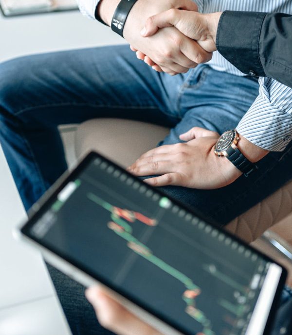 Business professionals shaking hands with a tablet displaying financial graphs.